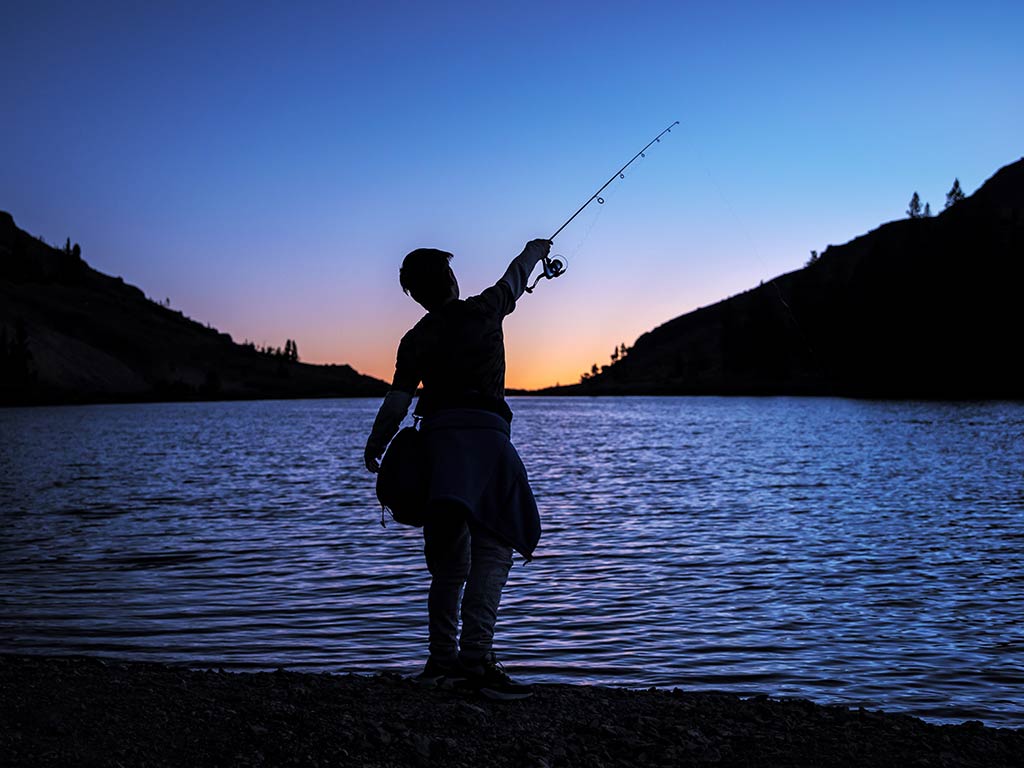 A view from behind of the silhouette of boy casting a fishing line into a lake in Nevada at sunset, with dark blue skies punctuated by a setting sun in the distance