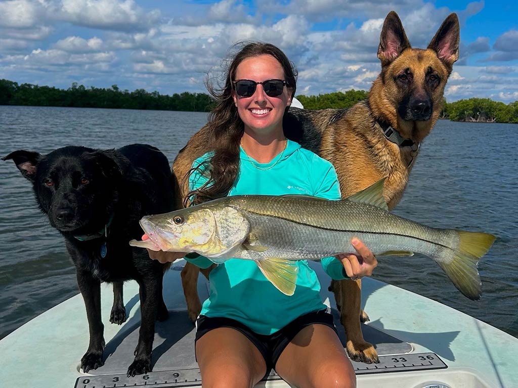 A woman in sunglasses sitting on the bow of a fishing boat and holding a Snook as two large dogs pose either side of her on a day with sunny intervals in Florida