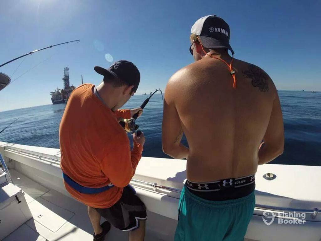 A view from behind of one shirtless man helping another who is struggling while bottom fishing next to an oil rig in the Gulf of Mexico on a clear day