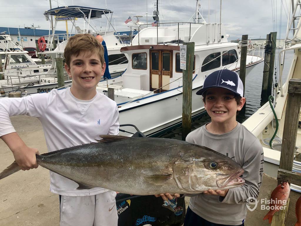 Two boys standing on a dock in front of some fishing boats and holding a sizeable Amberjack between them