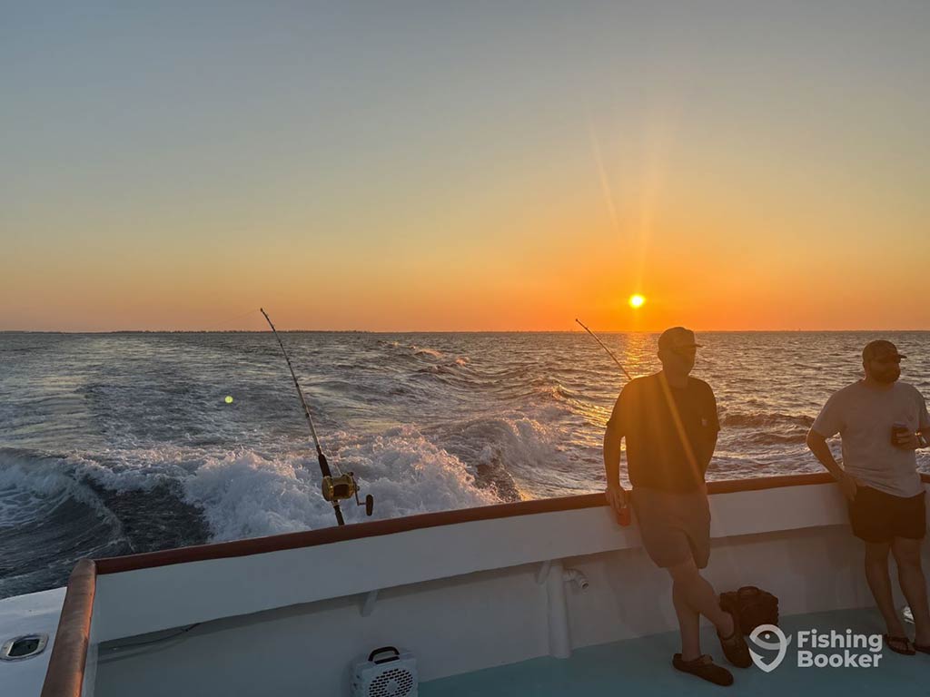 Two men leaning against the back of a deep sea fishing charter in Panama City, with two trolling rods visible behind them looking towards the sun setting in the distance across the water