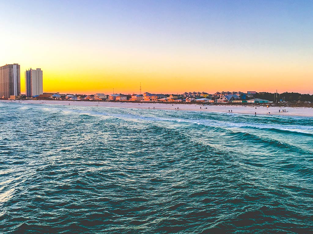 A view across some choppy waters towards the town of Panama City in Florida's Panhandle region at sunset, with an orange hue visible beyond a beach and two high-rise buildings in the distance