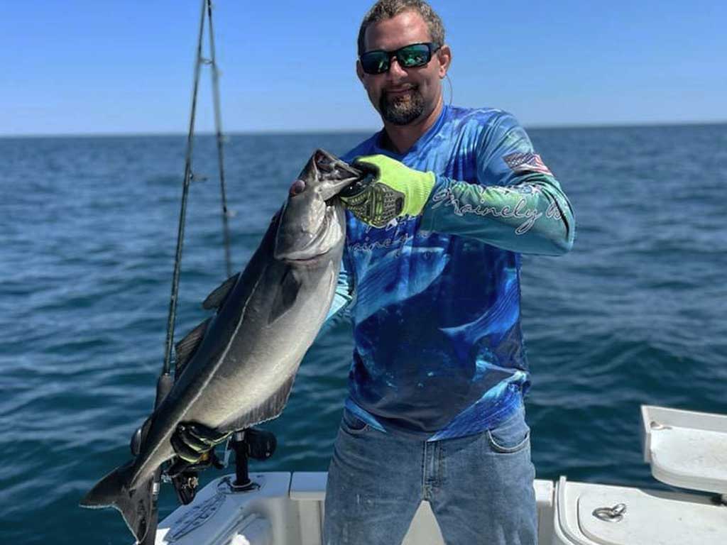 An angler standing on a charter fishing boat and posing with Pollock in both hands while looking directly at a camera on a bright and warm fall day