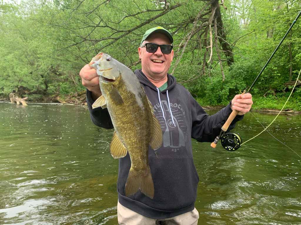 A smiling angler in sunglasses and a hat posing for a photo knee-deep in a river as he holds a Smallmouth Bass in one hand and his fly fishing rod in the other