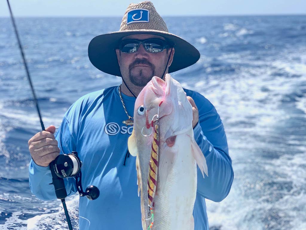 An angler in glasses and a hat standing on a boat, holding a rod in one hand and a Golden Tilefish in the other