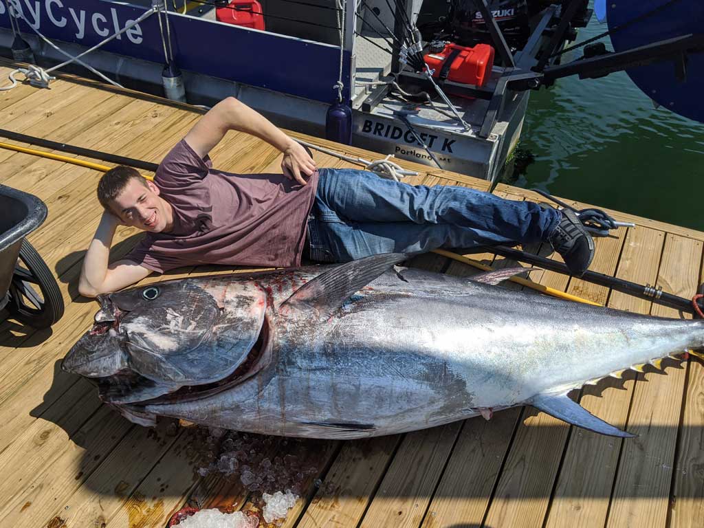 A photo of Tuna placed on the wooden deck floor after a deep sea fishing trip in Maine and an angler who caught it captured while posing on the floor next to Tuna