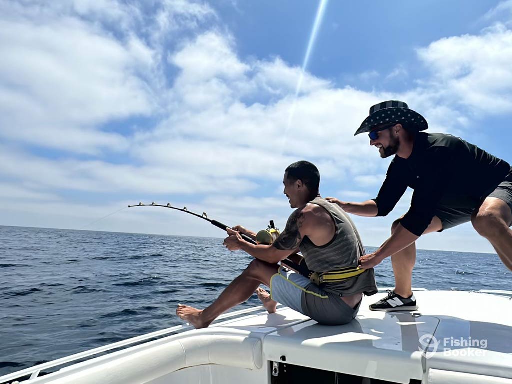 A man sitting on a fishing charter and bottom fishing in the Pacific Ocean, while another man attempts to distract him from behind on a clear day