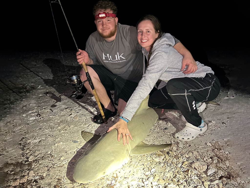 A man and woman crouching down on a beach at night, holding a Shark to the floor, as the man holds a fishing rod in his hand