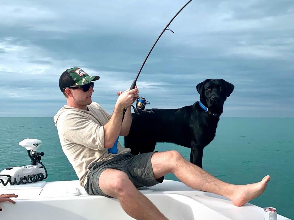 A man sitting on the bow of a fishing boat in shallow nearshore waters and struggling with a bent rod, as a black dog stands next to him, looking at the camera