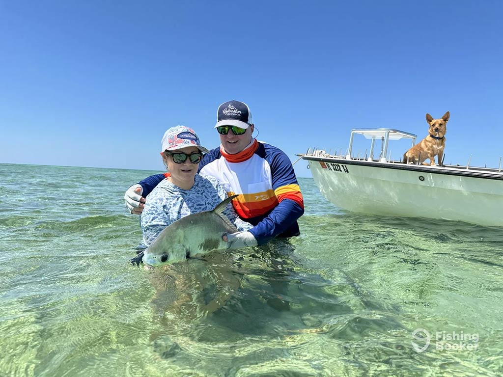 A man and woman stand waist-deep in crystal clear waters while releasing a Pompano fish, with a dog looking towards them from a bay boat behind them on a clear day