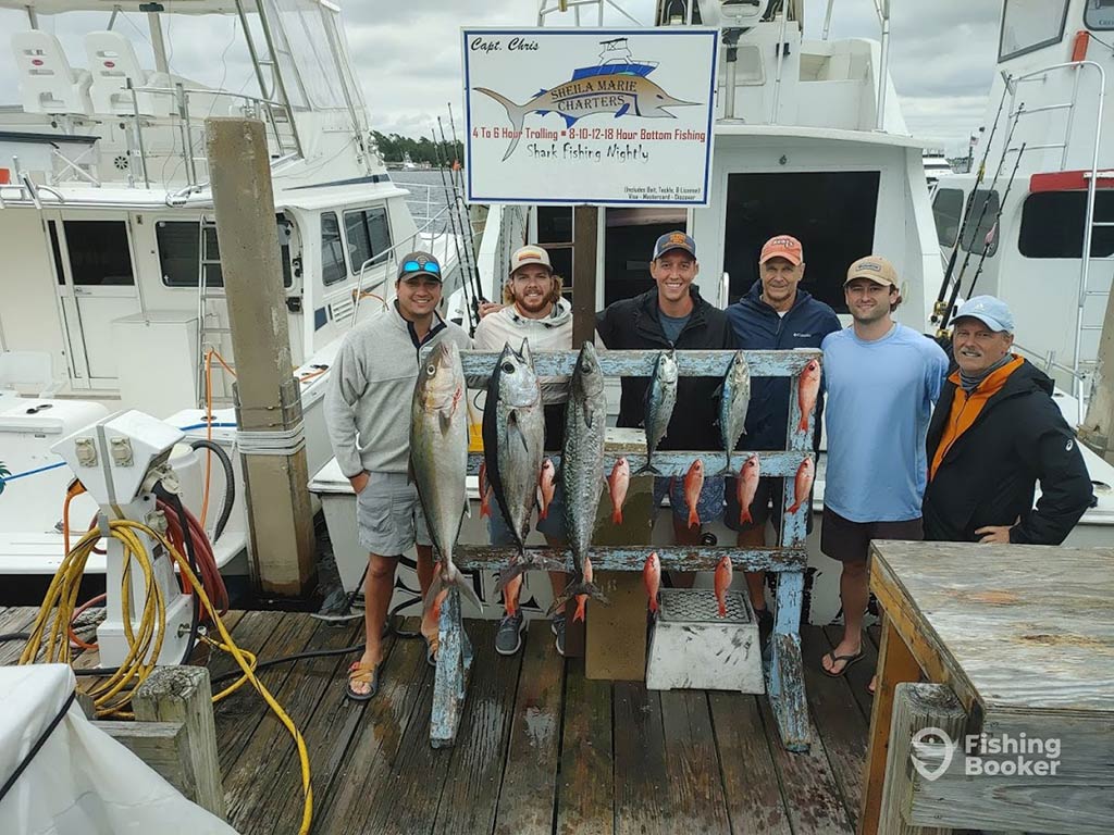 A group of male anglers showing off their catch on a deck in Panama City, FL, in front of their boat and a sign, with a mixture of Amberjack, Tuna, and Snappers hanging from a wooden sign