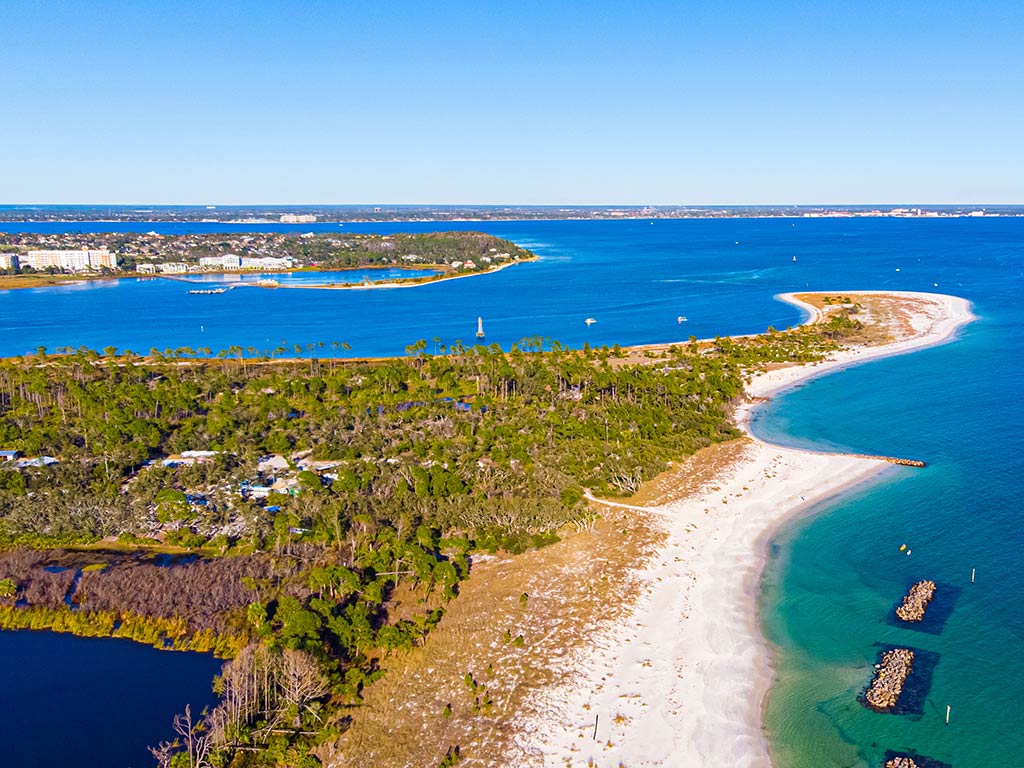 An aerial view of Panama City, Florida, on a clear day, with St. Andrews Bay visible in the distance, along with a small beach leading towards a jetty in the foreground