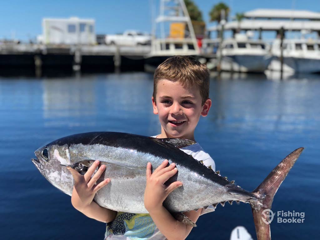 A young boy stands on a dock, holding a Blackfin Tuna with both hans in front of the bay's waters and a boat visible in the distance on a clear day