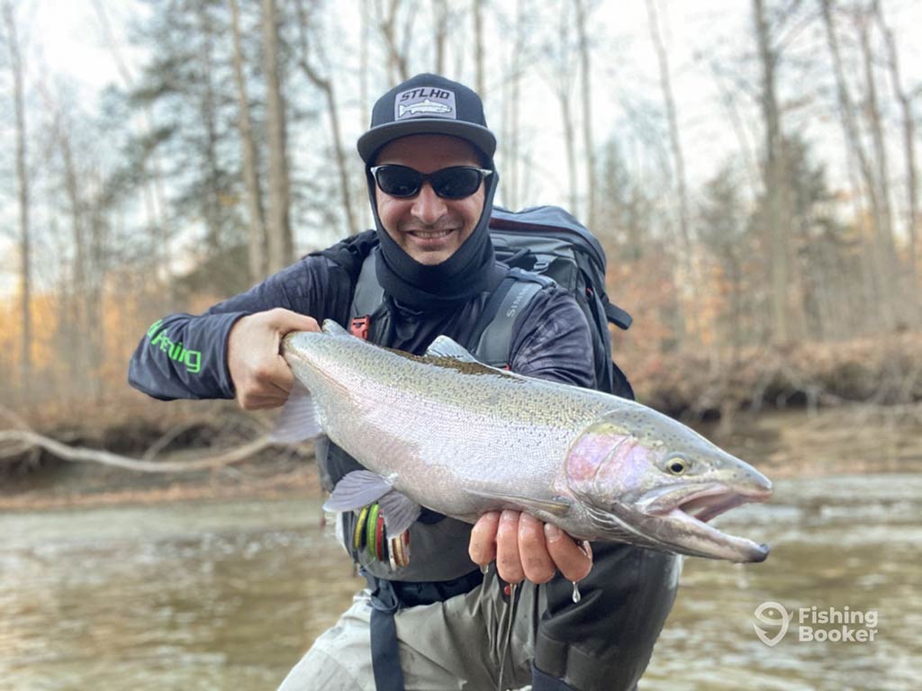 A man in a baseball cap and sunglasses crouching down and holding a sizeable Rainbow Trout towards the camera next to a river on a winter's day